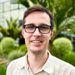 Photo of a male with glasses, with green vegetation and a tinted glass building in the background.