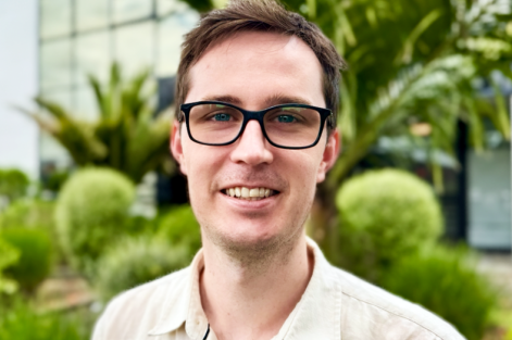 Photo of a male with glasses, with green vegetation and a tinted glass building in the background.
